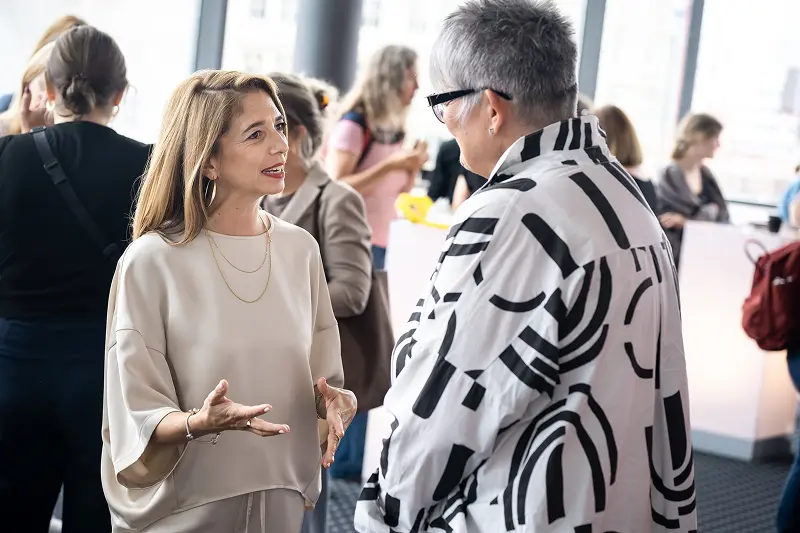 Bianca-Maria Klein und eine Kundin stehen bei einer Firmen-Veranstaltung im Rahmen ihrer Teambegleitung im Gespräch vertieft, wobei Bianca-Maria im hellen Oberteil gestikulierend die Bedeutung von klarem Erwartungsmanagement im Business-Kontext erklärt.