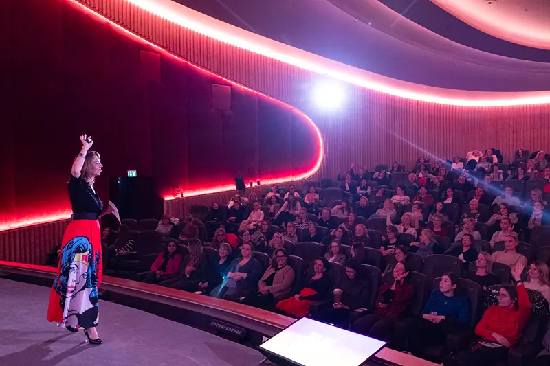 Bianca-Maria Klein in einem schwarzen Oberteil und einem auffälligen Comic-Print-Rock steht auf einer Bühne vor einem voll besetzten Saal und hebt motivierend die Hand, um die Bedeutung von klarem Erwartungsmanagement in ihrer Keynote bzw. ihrem Vortrag vor großem Publikum zu veranschaulichen.